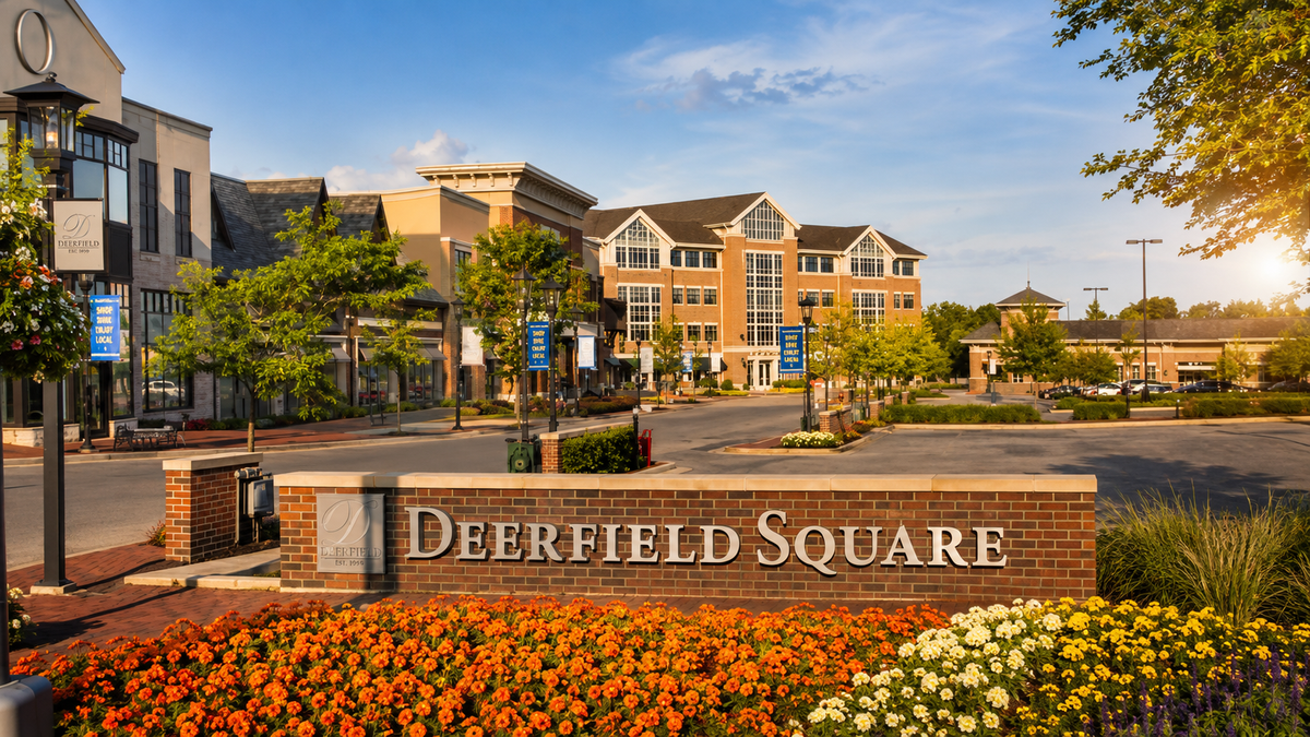 A brick sign reading "DEERFIELD SQUARE" in a landscaped plaza with orange and white flowers in the foreground, a shopping street with brick buildings, trees, and a blue sky in the background.