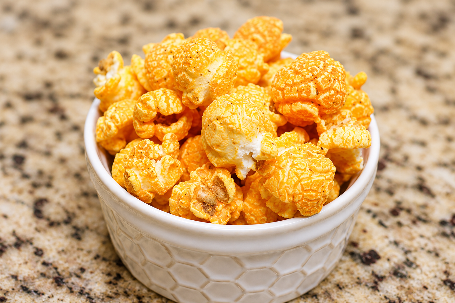 Crispy golden-orange popcorn piled in a small white ceramic bowl with a textured honeycomb pattern, set on a speckled beige granite countertop.