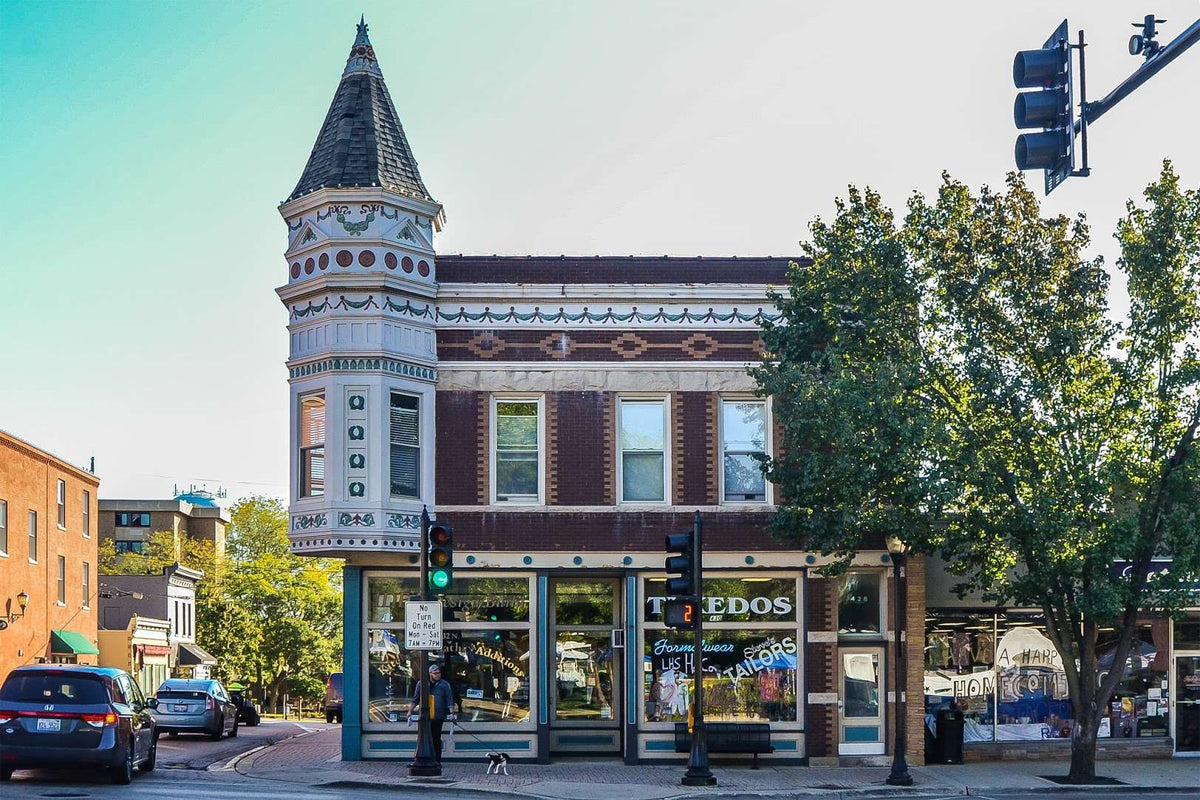 Libertyville, IL historic brick and white building with a turret and storefront, photographed on a city street with trees, cars, traffic lights, and a clear blue sky.