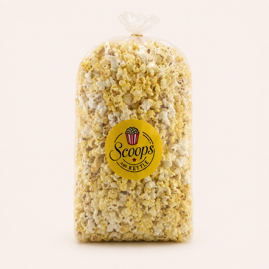 A clear plastic bag filled with popcorn, tied at the top, and featuring a round yellow label that reads 'Scoops of Kettle' on a plain light background.