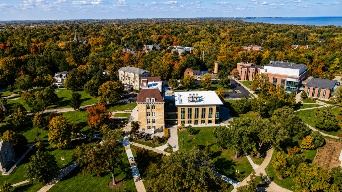 Aerial view of a campus with several brick and stone buildings surrounded by green lawns, trees, and pathways, with autumn foliage and a body of water in the distance.